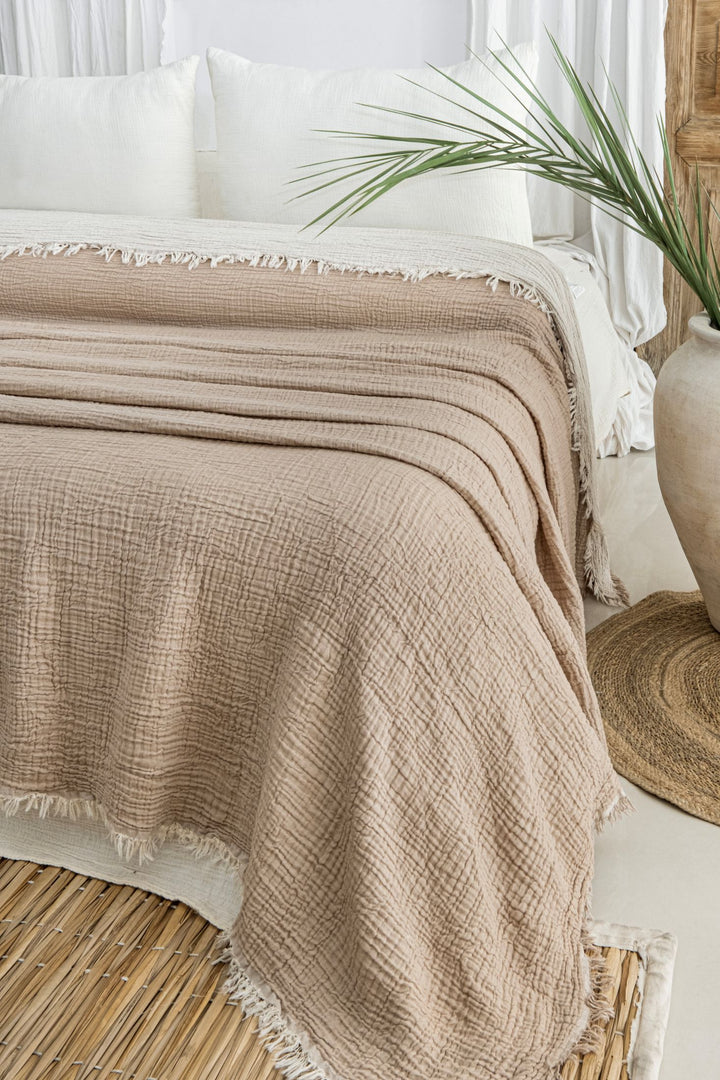 A neatly made bed features the Taupe Crinkled Muslin Bed Blanket by turkanhome.com with white pillows, beside a large vase with a green palm leaf, all set atop a natural woven rug in a bright, airy room.
