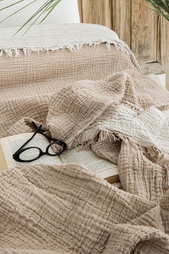 The Taupe Crinkled Muslin Bed Blanket from turkanhome.com is draped over an open book with black eyeglasses resting on the pages, while a wooden headboard and greenery in the background create a relaxing atmosphere.