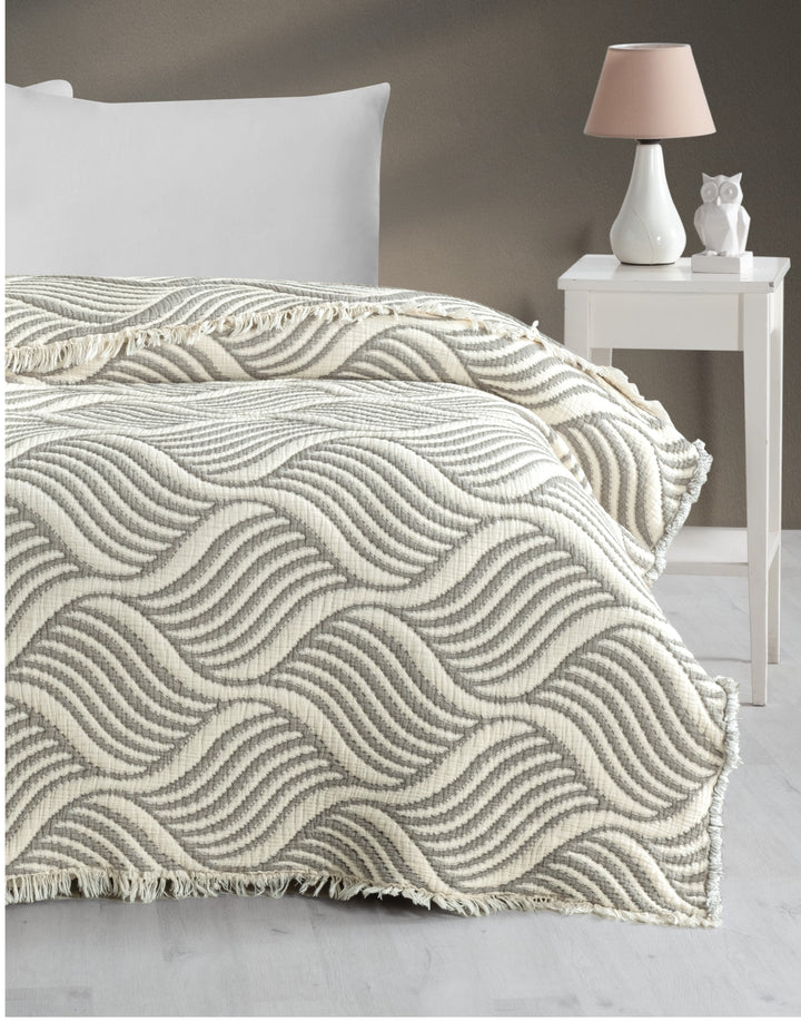 A cozy bed features the turkanhome.com Cozy Drift Muslin Quilt—a textured cream and gray bedspread with a wavy pattern—beside a white nightstand holding a beige lamp and an owl figurine, all set against a neutral wall.
