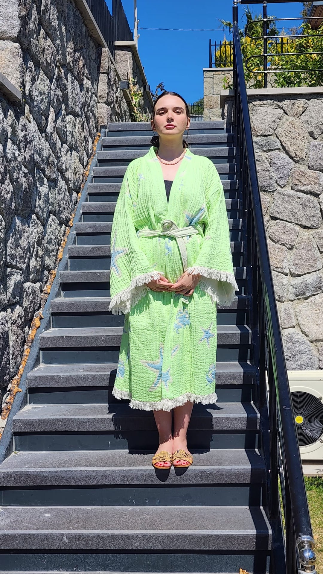 A woman stands on outdoor stone steps wearing the Green Crab Robe by turkanhome.com—styled as a kimono, lounge wear, or beach wear—with fringe and starfish patterns, paired with sandals and a topknot, facing the camera with hands folded.