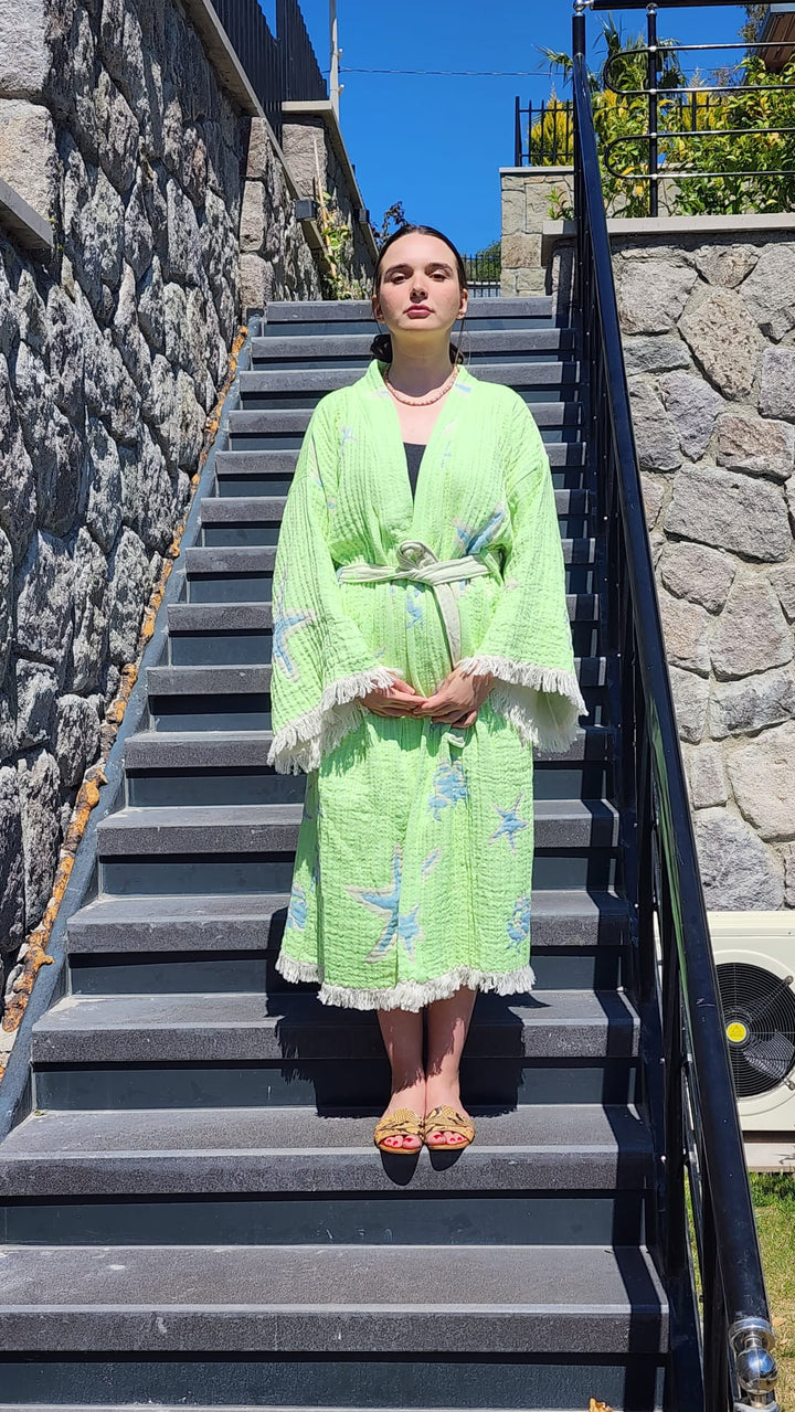 A woman stands on outdoor stone steps wearing the Green Crab Robe by turkanhome.com—styled as a kimono, lounge wear, or beach wear—with fringe and starfish patterns, paired with sandals and a topknot, facing the camera with hands folded.