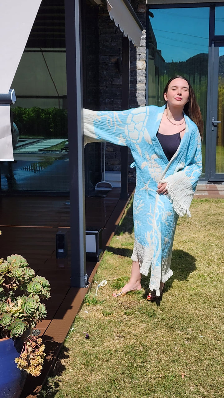 A young woman in the Blue Turtle Robe by turkanhome.com stands barefoot on grass by a modern building, holding a post. Sunlight highlights her as she looks upward, surrounded by greenery—perfect as kimono lounge or beach wear.