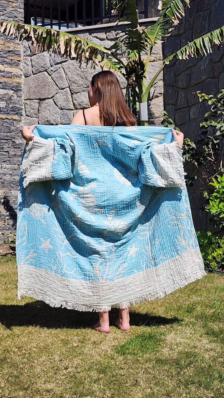 A woman stands barefoot on grass, back to the camera, holding open the Blue Turtle Robe by turkanhome.com—a light blue and white cotton muslin kimono with cloud and star designs—outdoors by a stone wall and tropical plants.