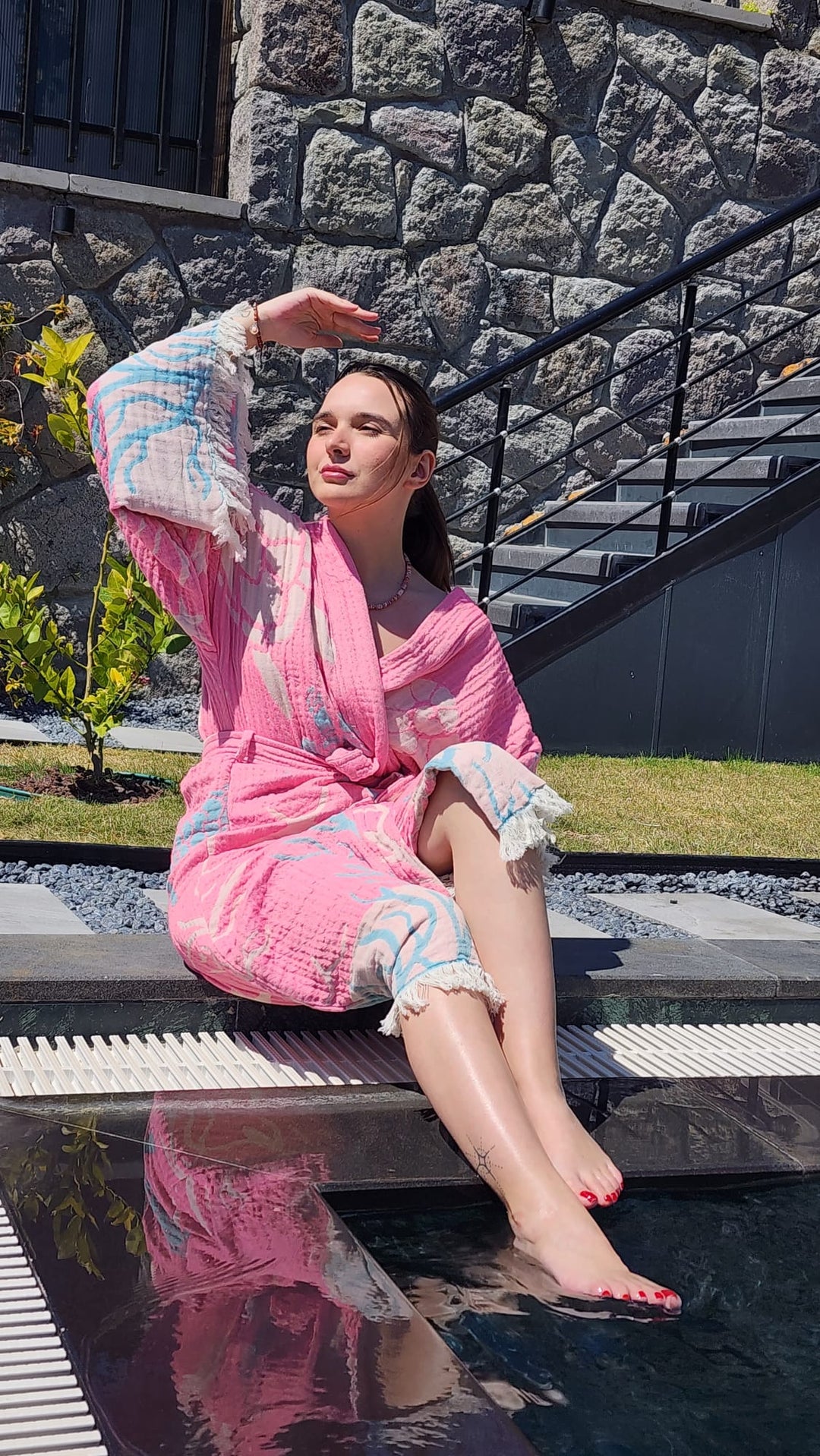 A woman lounges by the pool in a Pink Turtle Robe from turkanhome.com—crafted from 100% natural cotton for kimono-style comfort—one leg in the water as she shades her eyes, with lush greenery and a stone wall behind her.