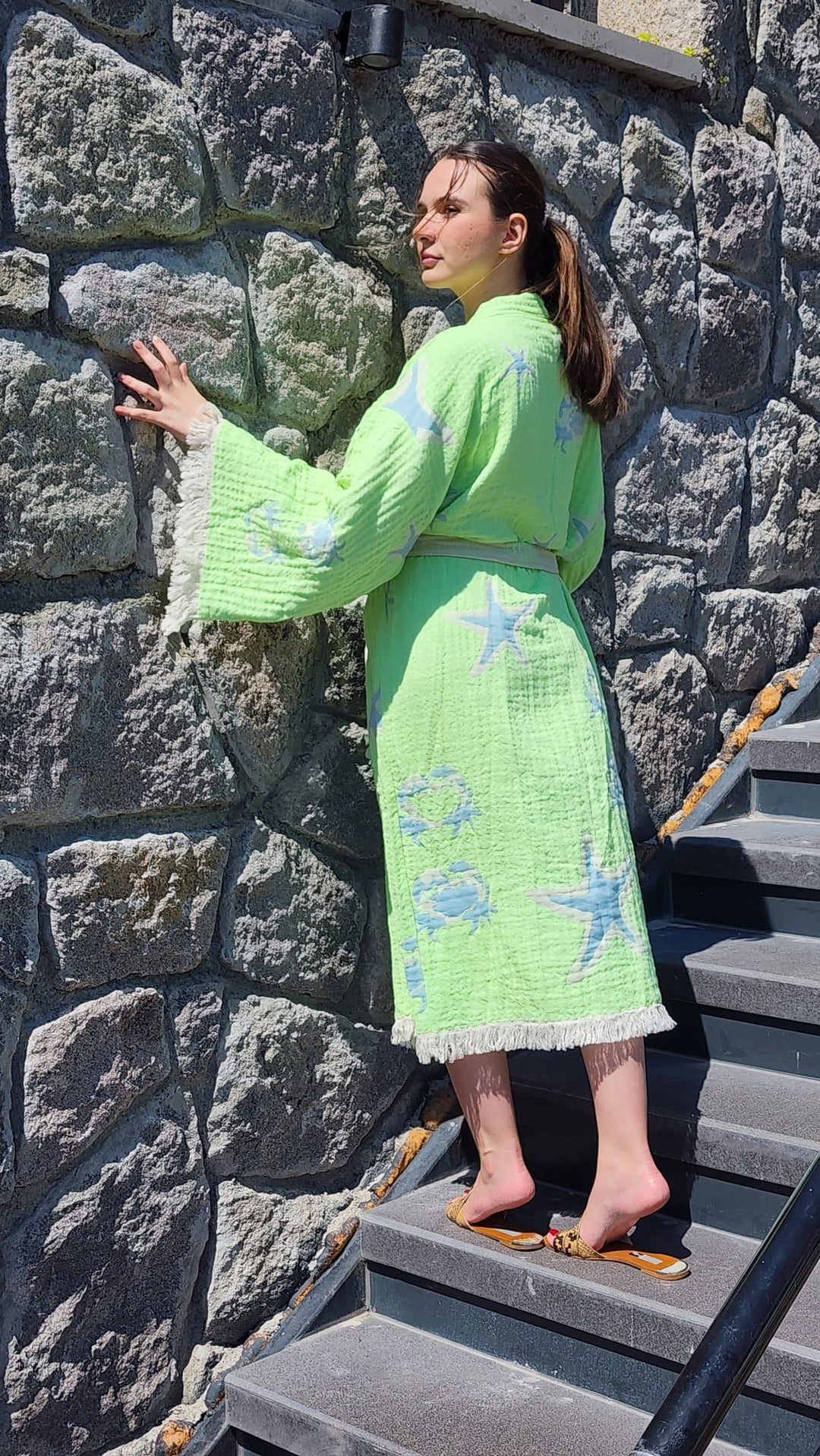 A woman in the Green Crab Robe kimono by turkanhome.com stands on outdoor steps, touching a stone wall and looking to the side in bright sunlight.