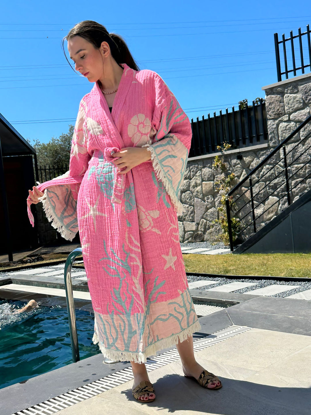 A woman in a Pink Turtle Robe by turkanhome.com, a 100% cotton kimono for lounge or beach wear, stands by a pool on a sunny day, looking down and holding the belt. Stone walls and metal railings are in the background.