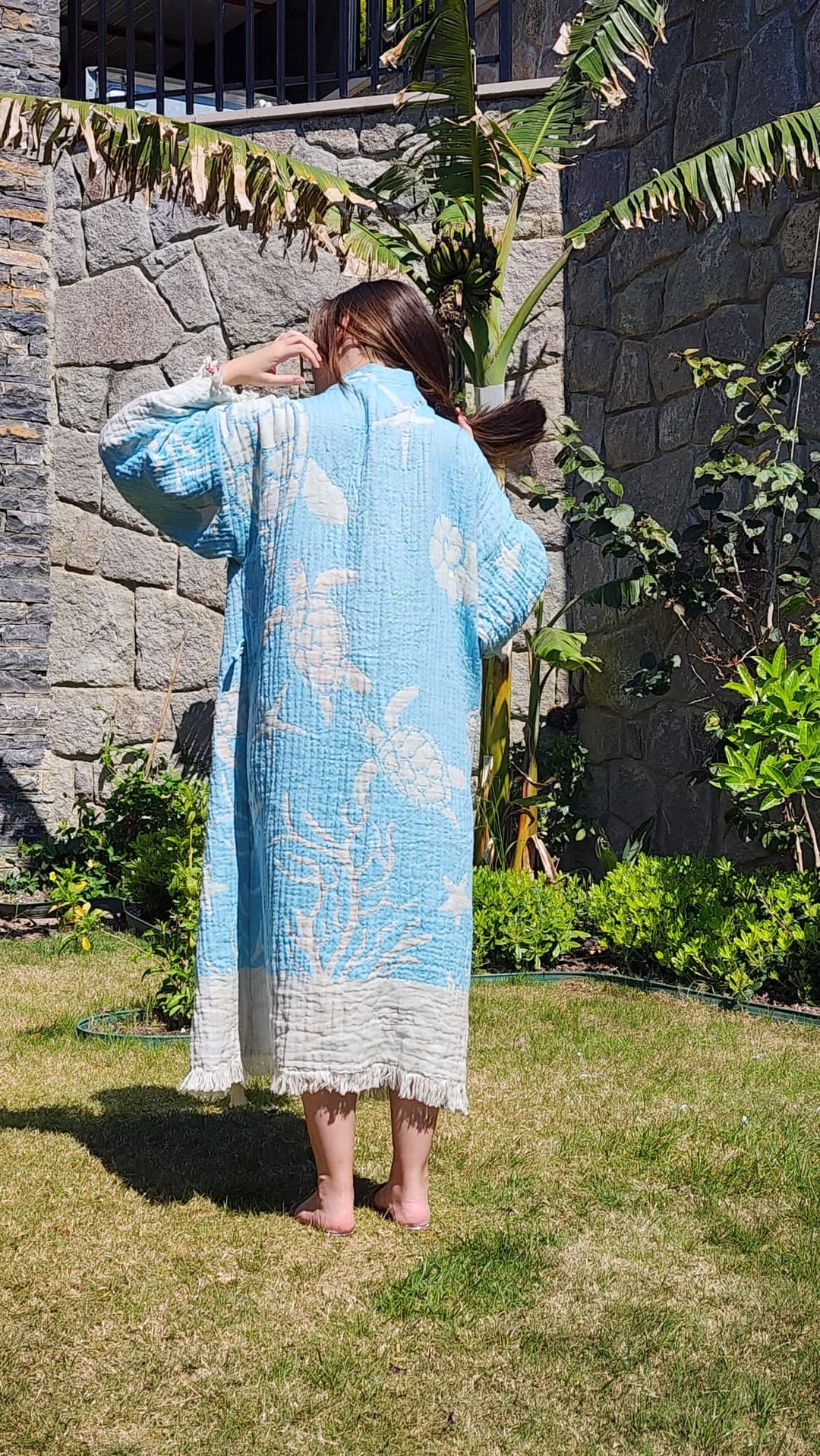 A person with long brown hair stands barefoot on grass, facing away, wearing the Blue Turtle Robe from turkanhome.com. The light blue kimono features white ocean-themed turtle patterns. Stone walls, green plants, and a banana tree are behind them.