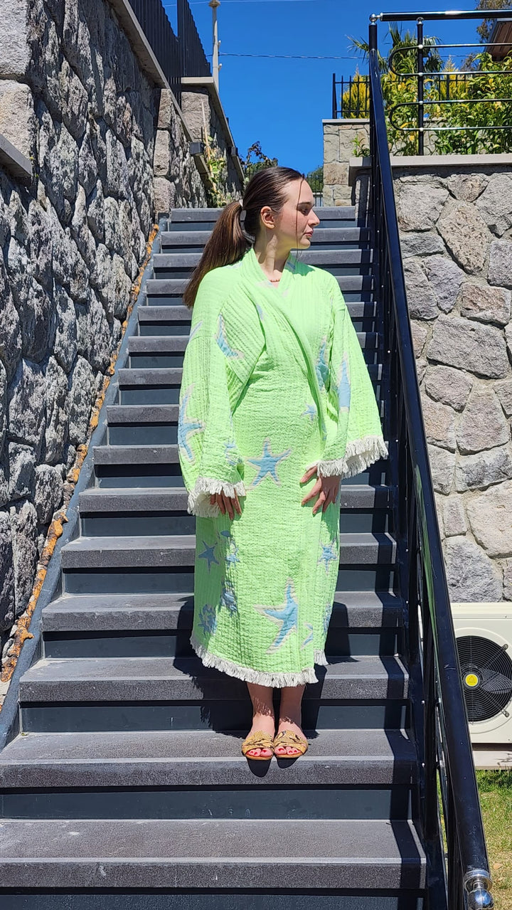 A woman wears the Green Crab Robe—a light green, patterned natural cotton kimono with fringes from turkanhome.com—as she stands barefoot atop stone steps outside, framed by a stone wall and a clear blue sky.